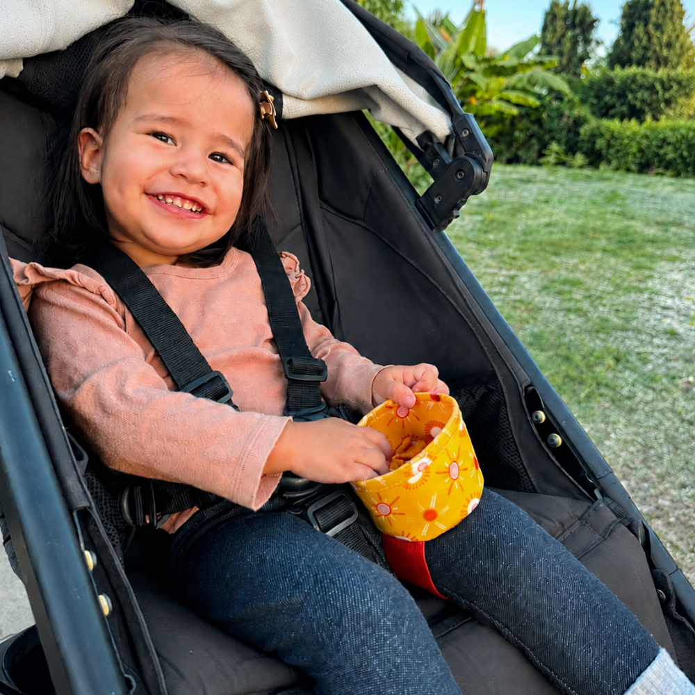 A young child sitting, buckled in a stroller outdoors, holding a yellow sun patterned wearable snack cup comfortably strapped around her seated thigh. The child is wearing a dusty pink long-sleeve top and dark blue jeans, secured in a black stroller with a cream canopy.